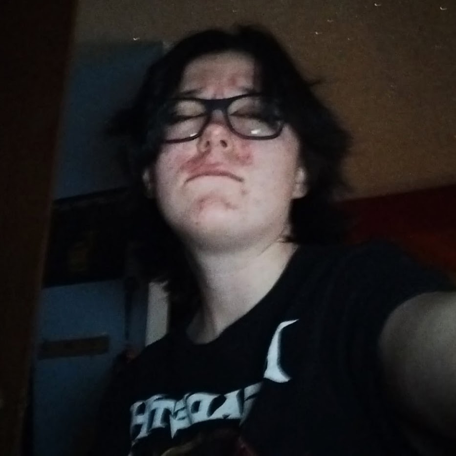 selfie of a young white person with medium-length dark brown hair, black glasses, and a Black megadeth t-shirt closing their eyes and biting their bottom lip goofily in a dark bedroom